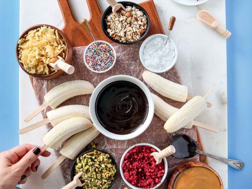 A wooden board placed on top of a piece of marble. Displayed on the board are frozen bananas and assorted gluten-free and vegan toppings to make frozen vegan banana pops including gluten-free sprinkles, gluten-free pretzels and crushed nuts.
