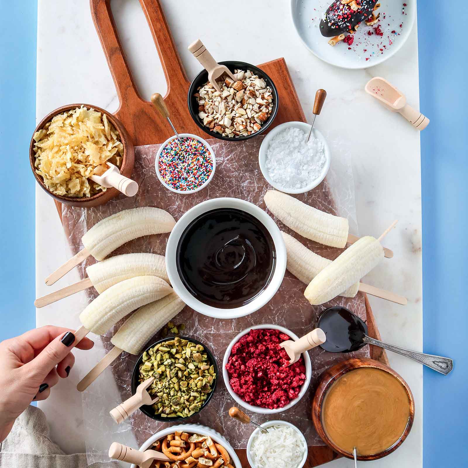 A wooden board placed on top of a piece of marble. Displayed on the board are frozen bananas and assorted gluten-free and vegan toppings to make frozen vegan banana pops including gluten-free sprinkles, gluten-free pretzels and crushed nuts.