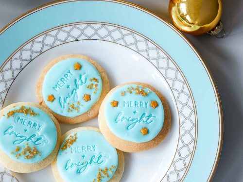 Merry and Bright Gluten-Free Sugar Cookies with blue fondant and gold sprinkles and stars arranged on a blue and gold plate with Christmas decorations surrounding the plate.
