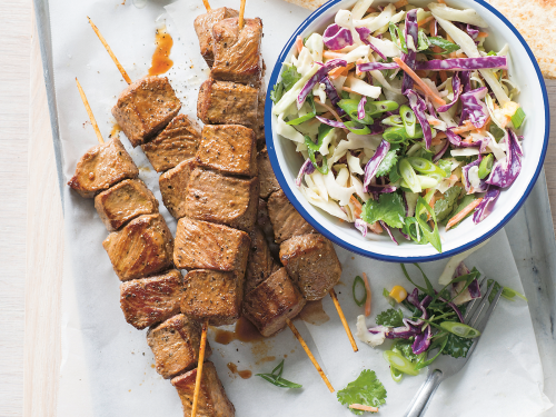4-ingredient-lamb skewers arranged on a baking tray lined with baking paper. A white bowl with blue rim sits to the right full of fresh slaw. Lime wedged and corriander have been arranged on the baking tray and gluten-free tortillas sit just at the top right hand side of image.