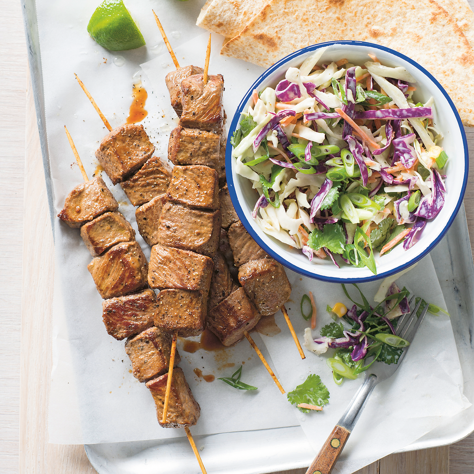 4-ingredient-lamb skewers arranged on a baking tray lined with baking paper. A white bowl with blue rim sits to the right full of fresh slaw. Lime wedged and corriander have been arranged on the baking tray and gluten-free tortillas sit just at the top right hand side of image.