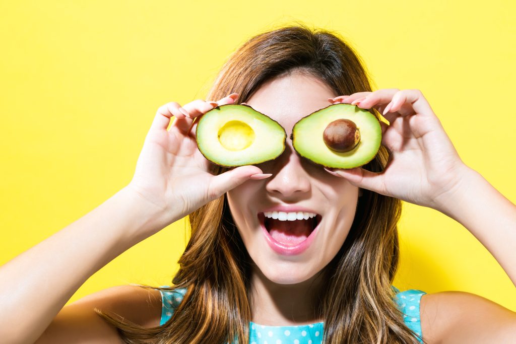 Happy young woman holding avocado halves on a yellow background.