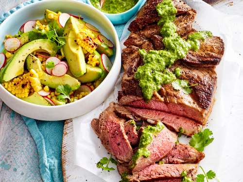 BBQ Lamb sits on greaseproof paper on a round white cutting board. Half of the lamb is sliced ready to serve. The lamb is topped with a drizzle of Avocado Chimichurri Dressing. A white bowl of corn, avocado and radish salad sits to one side and at the back is a blue bowl with extra avocado chimichuri.