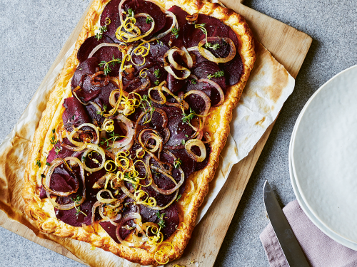 Beetroot, onion and thyme galette sitting on a wooden chopping board. A knife sits on a dusty pink napkin ready to cut and serve.