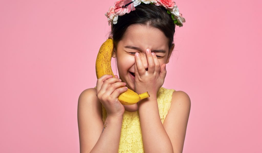 Happy girl holding banana on a pink background.