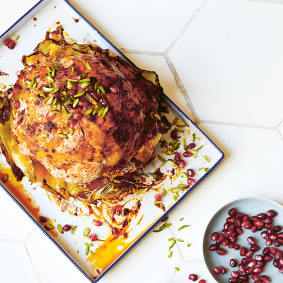 Harissa-yoghurt whole roasted cauliflower sits on a baking tray. A small blue bowl with pomegranates sits to the side.