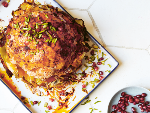 Harissa-yoghurt whole roasted cauliflower sits on a baking tray. A small blue bowl with pomegranates sits to the side.