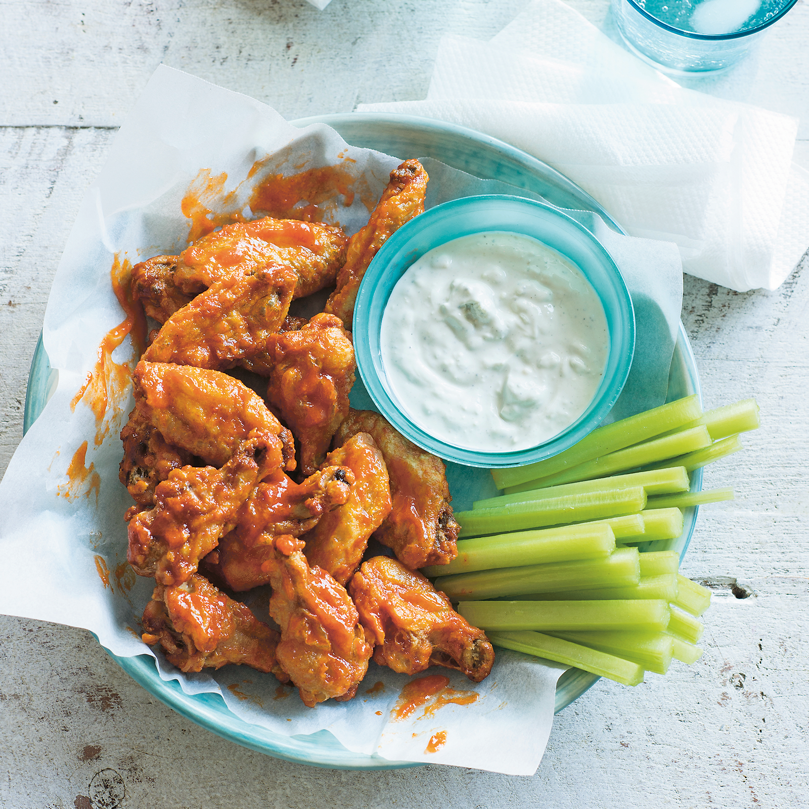 Gluten-free baked buffalo wings in a large bowl lined with greaseproof paper. A small dish with blue cheese dipping sauce is nestled next to the wings alongside celery sticks.