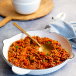 A white cast-iron frying pan filled with savoury mince with carrots and garnishd with fresh parsley. A gold serving spoon is placed in the mince ready to serve into bowls resting to the side;