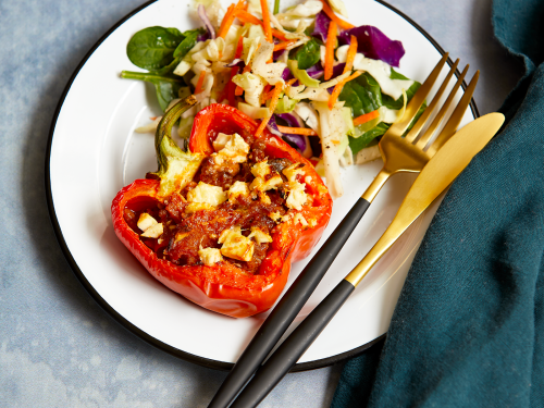 Half a red capsicum, cut in half vertically. Capsicum is stuffed with savoury mince and feta. It sits on a white enamel plate with a black edge with a side salad and black and gold cutlery.