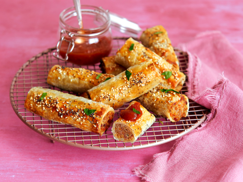 Gluten-free family favourite sausage rolls arranged on a rose gold wire rack with a glass jar of tomato sauce. The rack sits on a pink background and a pink napkin rests on the side.