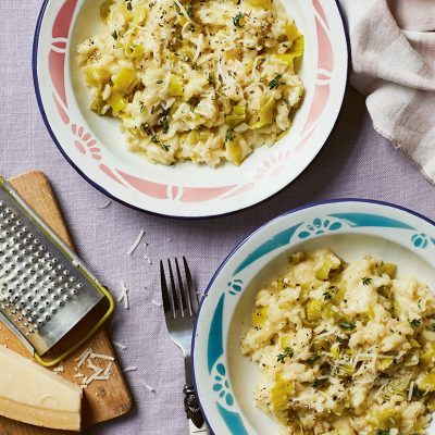 Two bowls of Cheesy Leek, Thyme & Taleggio Risotto prepared in a slow cooker. To the side of the bowls is a wooden chopping board with a block of Parmesan and a grated. The risotto has been sprinkled with freshly grated Parmesan.