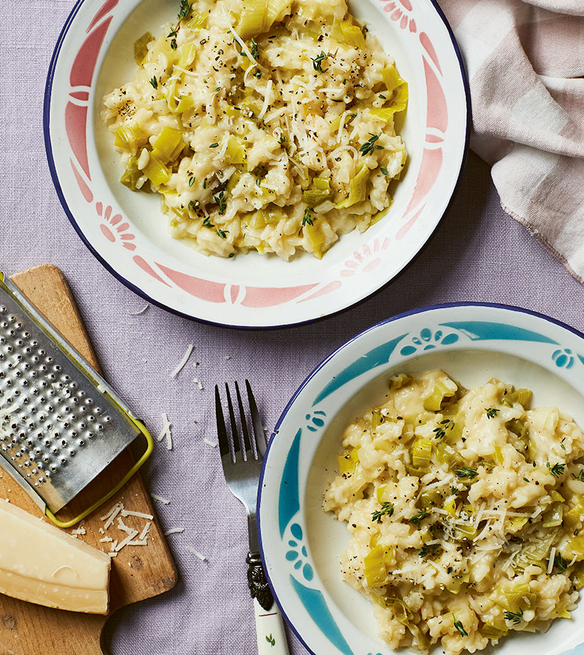 Two bowls of Cheesy Leek, Thyme & Taleggio Risotto prepared in a slow cooker. To the side of the bowls is a wooden chopping board with a block of Parmesan and a grated. The risotto has been sprinkled with freshly grated Parmesan.
