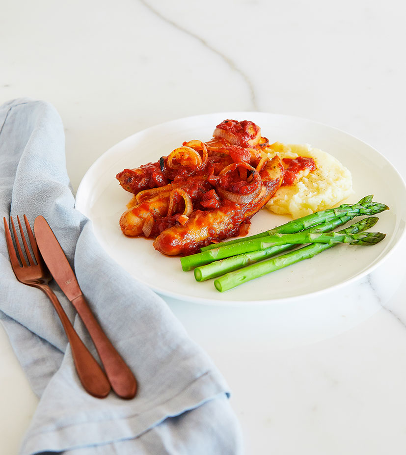 Slow cooker devilled sausages resting on a bed of mashed potato next to bright green steamed asparagus spears. This meal is free-from gluten and dairy.