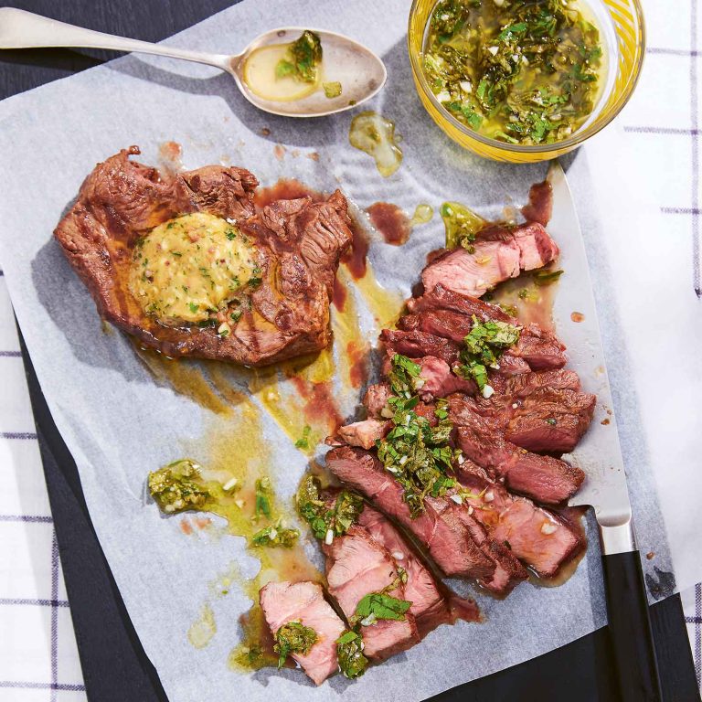 Credit-crunch flat-iron steak served on parchment paper, with one steak sliced and garnished with herbs and a whole piece topped with a dollop of butter, accompanied by a bowl of herb sauce.